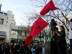 Three red flags with IWW logos being held above a crowd of people.