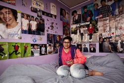 A teenage girl sits on a large bed with a grey blanket in her room, which has purple walls. The walls are adorned with posters of a boy band.