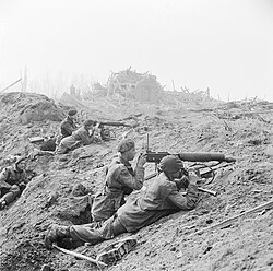 Five men with two guns in a devastated landscape. The remains of a bombed town are in the background