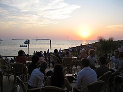 The upper half of the photograph is a sky with some clouds. The shape of the sun is not distinct but it is several degrees over the horizon. The lower half of the photograph shows the calm sea with some recreational vessels. On the shore, there are the silhouettes of tens of people sitting at tables with drinks. Most of the people look towards the sun. Among the people, there are some tiki torches and plants.