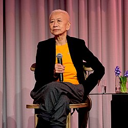 Shu Lea Cheang sitting in a chair on a stage, holding a microphone, looking into the audience