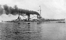 A large warship plows through the water, thick black smoke pouring from its two central smoke stacks.