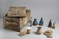 A painted wooden box with lid askew and its contents of stone and glass vessels neatly arranged beside it