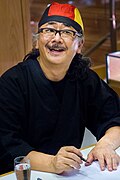 Portrait of a mustached Japanese man wearing glasses and a black, red, and yellow head bandana.