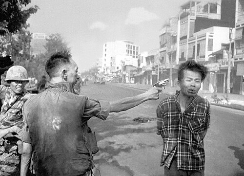 Nguyễn Ngọc Loan aims a pistol at Nguyễn Văn Lém. Lém's face is visibly distorted from a bullet that is still travelling through his head.