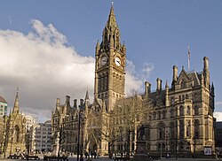 A grand gothic revival building as viewed from afar, with a gothic revival clock tower stretching high into the sky. Trees partially obstruct the view and the sky is clear with some clouds.