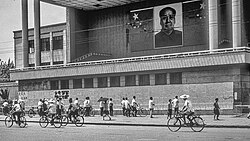 A group of pedestrians and cyclists by a large poster with Chinese political writing, alongside a portrait of Mao Zedong and a Chinese flag