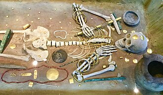 Skeleton in a museum case, with many golden offerings covering and surrounding the body
