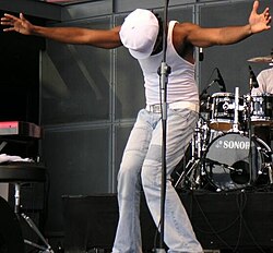 A man wearing white pants, white undershirt, and white hat standing on stage with his head down and his arms open wide.