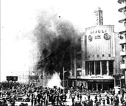 A crowd of bystanders stands outside a burning building, with black smoke coming out of the windows.