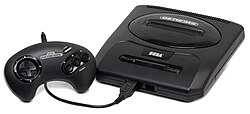 Photograph of an electronic video game console with single controller plugged in the front. The console is encased in black matte plastic that is shaped as a flattened rectangular cuboid with rounded corners. The top has two buttons and a slot for cartridges, while the front has two ports. A black game controller with a black cable is plugged into one of the ports.