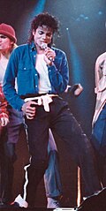 Photograph of a man performing onstage, wearing a blue shirt and black pants while holding a microphone, with backup dancers visible behind him.