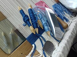 Tapestry tools, on the loom. Bobbins, scrapper (with short teeth), comb (double-ended), and awl (tip hidden).