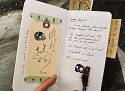 A journal held open by a barely visible hand. The left page has a drawing of a pronghorned kangaroo rat, a skull and a cactus taped to the page with washi tape; the vertical caption states "cacti and vertebrae". The right page has a handwritten journal entry with another skull drawn under it as well as a physical feather; at this resolution, the text is mostly illegible.