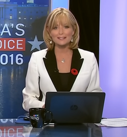 Woman with blonde hair, sitting at a desk with a laptop computer, wearing a white suit coat, and a black shirt with a red poppy on the lapel