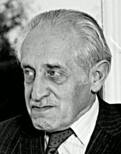 Lean-faced man in striped suit, aged around 80, with moustache and white hair combed back 1940s-style.
