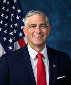 Official House portrait of Baumgartner smiling in front of the U.S. flag, wearing a black suit with American flag lapel pin, white shirt, and red tie.