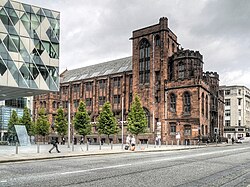 a brick building in the background as viewed from across a road, made from one 3-storey section of gable (normal triangular) roofing, then a tall 4-storey tower as the next part of the building to its right, than a smaller, 2-storey tower on the very right.