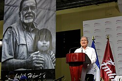 A photograph of Salvador Sánchez Cerén in front of a mural of Schafik Hándal