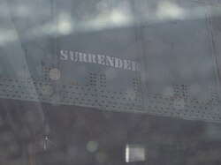 Miniature SURRENDER graffiti from 2007 stenciled on the green railroad bridge. Above the stencil are dark green squares where the previous graffiti has been painted over.