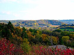 Landscape with low rolling hills, farm buildings in the middle distance surrounded by fields, and bright early autumn foliage