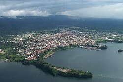 In the distance are cloud covered mountains, while in the front of the picture are houses leading to the harbour which has two piers leading out to sea