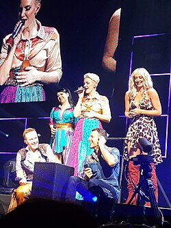 A group of five people – three women (pictured standing) and two men (pictured crouching) – standing on a stage performing a music act.