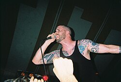 Vocalist Andrew Schwab performs at a concert. He has shaved his head and is wearing a black sleeveless shirt that displays his tattoos.