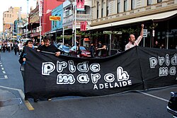 People on street holding a black sign with silver lettering that says "Pride March Adelaide".