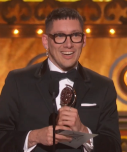 Headshot of Scutt in a tuxedo against a golden background, clutching an award