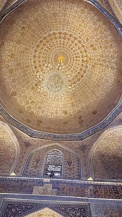 Photograph from the ground of a round dome. The lower stage is square. The stage under the dome is octagonal and has a window visible in the photograph. The dome is covered in symmetric golden patterns resembling a fleur-de-lys. The patterns are set in circles encircling the dome, smaller and smaller towards the center. An unlit chandelier hangs from the top of the dome.