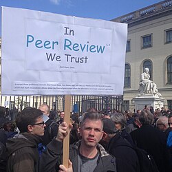 A protestor in a crowd holds up a sign reading, in the Comic Sans font: In Peer Review [(footnote 1 and 2)] We Trust / (and Comic Sans). Below, footnote 1 reads "except those predatory journals. Don't trust those. See Nature 543, 481–483 (23 March 2017) doi:10.1038/5434812"; footnote 2 reads "let's not start an academic debate about the flaws of peer review when the alternative is to trust someone's gut feeling"