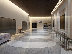 View of the lobby, facing north, along the passageway on the western side of the elevator core. There are white and gray pavers on the floor. To the right is a wall with the artwork "Portals", which includes alternating bands of white and brown glass surrounding a set of bronze and nickel plates.