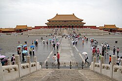 A large courtyard: in the foreground are steps with the photographer standing on them; beyond lies the courtyard with groups of tourists standing and walking, and in the background, on a raised terrace, stands a palace building
