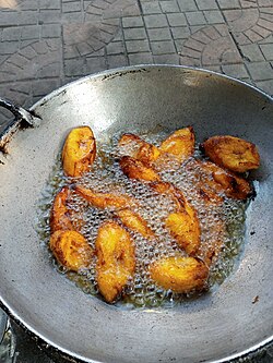 Slices of plantain being fried in a wide pan outside in bubbling oil