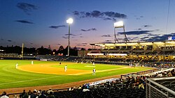 A view of the green baseball field from the third base line seats showing men in white baseball uniforms playing their positions as the sun has just set behind first base