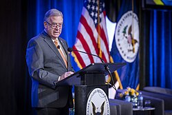 A man with glasses and a suit speaks at a podium