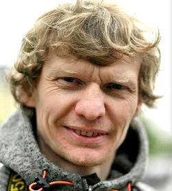 Colour press photo of smiling, clean-shaven man with wavy blond hair.