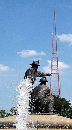 The tall, red tower against a blue sky at day with a fountain and sculpture in the foreground.
