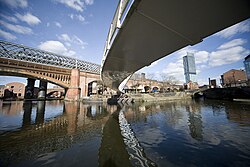 A photo taken from underneath a modern bridge over a river, with the underside of the bridge curving from the top right to the centre to the centre right of the frame. In the background is a grand railway bridge approximately perpendicular to the viewer which the first bridge then becomes parallel to the right of, and there is a skyscraper on the centre right as well.