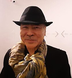 Portrait of a Japanese man wearing a black fedora hat, a black jacket, and a yellow and brown scarf.