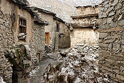 A corpsman in desert fatigues stands in the middle of a group of very old houses.