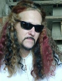 Three-quarter profile colour publicity shot of a man wearing sunglasses, with serious expression, minimal moustache-beard combo, and cascades of long, wavy brown, red, and black hair.