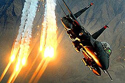 An F-15E Strike Eagle releasing a stream of bright flares over mountainous terrain in Afghanistan