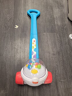 A photograph of a pre-school age girl in a raincoat walking in grass while pushing a ride-along Corn Popper by the handle.