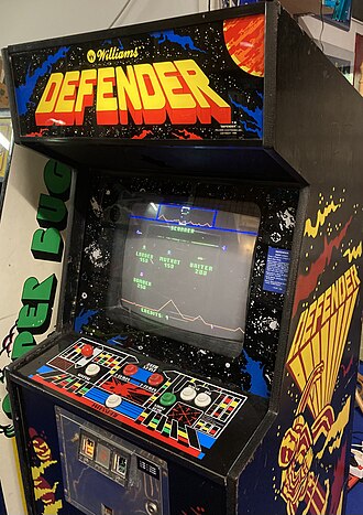 A vertical wooden structure with a visual display unit embedded in the front side. The sides have black panels with yellow and red artwork that reads "Defender", and the top front panel is a backlit marque that reads: "Williams. Defender". A red joystick with a groups of buttons are below the monitor, which displays a pixelated game screen.