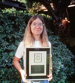 A woman standing outdoors in front of a shrub wearing a t-shirt, jeans and glasses. She is holding a plaque that features a one-color rendition of the cover art and states "River Raid - Presented to Carol Shaw in recognition of sales in excess of 500,000 units - January 25, 1983".