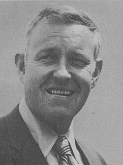 A bust-length black and white photograph of man with short dark-grey hair wearing a suit and tie. His mouth is open and his head and eyes are turned slightly to his right.