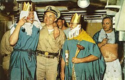 Color photograph of crossing the line ceremony held in the crews mess compartment of the nuclear submarine USS Triton that includes (from left to right) crewman Wilmot A. Jones dressed as the Queen of the Royal Court; Captain Edward L. Beach dressed in khaki uniform and ceremonial sword; Chief Loyd Garlock dressed as King Neptune, Ruler of the Raging Main; crewman Ross MacGregor dressed as Davy Jones in background; and crewman Harry Olsen dressed as the Royal Baby.