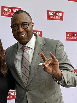 Howell in a gray suit smiling, making a wolfpack hand sign